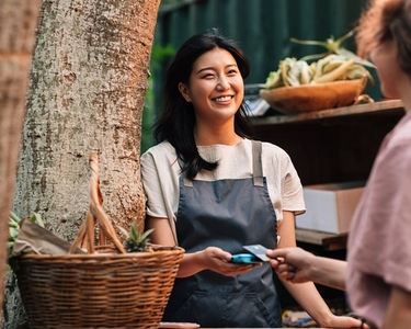 Happy Asian woman in an apron receiving payment on the outdoor market  Vendor with a card machine smiling and looking at the customer
