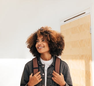 Girl with backpack looking up while standing in a corridor  Smiling female student with curly hair