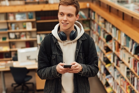 Male student with smartphone standing in library