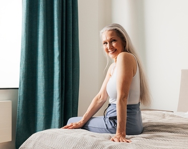 Smiling senior woman with long white hair looking at the camera while sitting on a bed  Female in casuals in bedroom