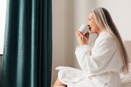 Senior woman drinking coffee in morning in bedroom  Female in bathrobe sitting on a bed looking at window