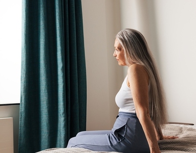Side view of an aged woman with a long white hair sitting on a bed looking at window