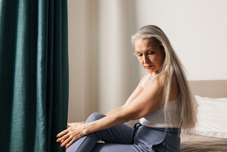 Sad and thoughtful senior woman sitting in bedroom and looking down