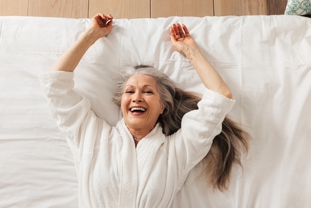 Happy senior woman in a bathrobe lying on a bed in a hotel room looking at camera