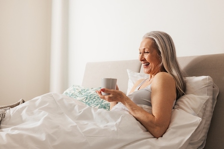 Laughing woman with a cup in bed  Smiling aged female holding a cup in bedroom