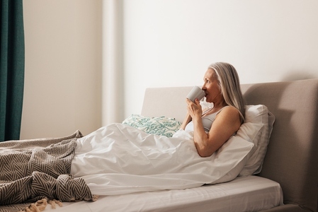 Senior woman drinking from a cup while lying in bed at home