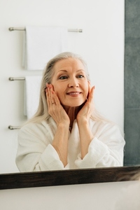 Senior woman with white hair touching her face with hands looking at a mirror in bathroom