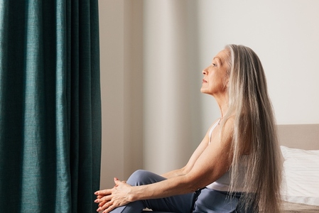 Pensive senior female with long hair sitting on a bed looking at a window