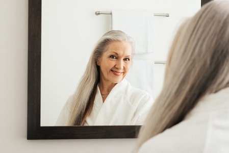 Older woman with long beautiful hair admiring herself in a mirror  Senior female in a bathroom in morning