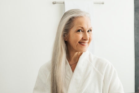 Smiling woman with long white hair at morning in bathroom  Aged female in bathrobe looking at her reflection in the mirror