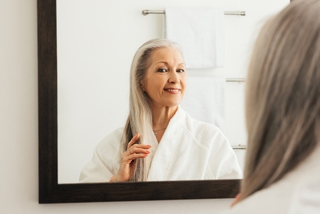 Aged woman admires her long gray hair in bathroom mirror