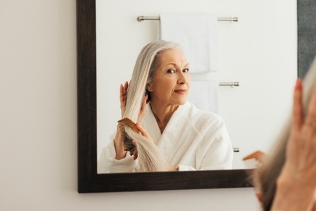 Senior woman looking at mirror while combing her long hair in the bathroom