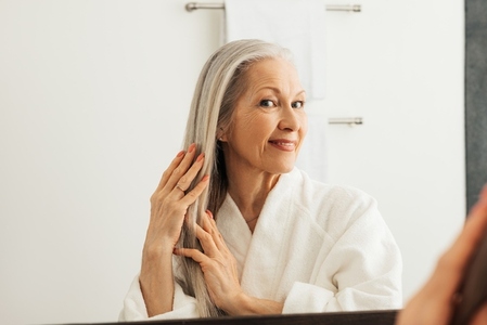 Woman with grey hair in the bathroom in front of a mirror  Senior female adjusting long hair with hands