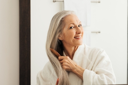 Smiling woman combing her long gray hair in front of a mirror