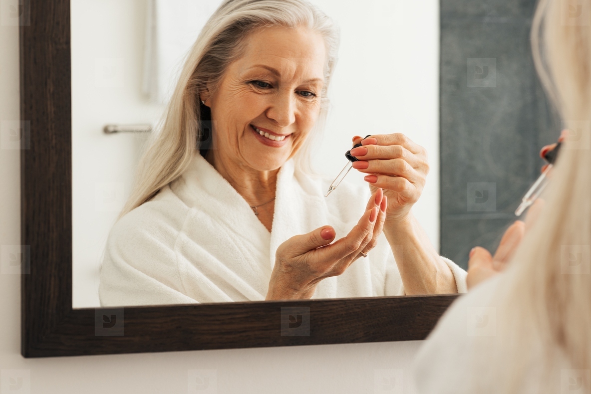Reflection in mirror of a smiling aged woman with a pipette. Smiling senior female in a bathroom using liquid face treatment