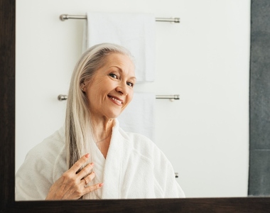 Smiling aged woman touching her long gray hair looking at her reflection in the bathroom mirror