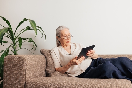 Senior woman in glasses lying on a sofa holding a digital tablet  Aged female in casuals reading from the portable computer