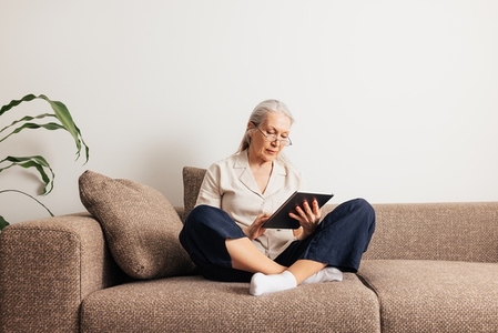 Senior woman sitting on a sofa with crossed legs holding digital tablet  Aged female reading from a portable computer at home