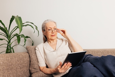Aged woman in eyeglasses lying on sofa and reading from digital tablet