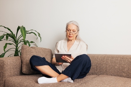 Aged woman in eyeglasses sitting on a sofa holding a digital tablet