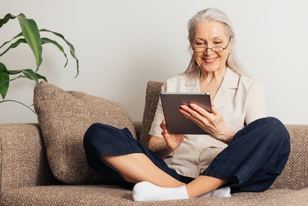 Smiling senior woman sitting with crossed legs using a digital tablet  Aged female in eyeglasses reading from a digital tablet at home