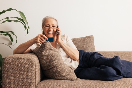 Happy senior woman holding a credit card and making call  Aged woman in glasses lying on sofa and shopping online