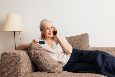 Smiling senior woman making a call via mobile phone and holding a credit card while lying on sofa