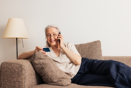 Senior woman looking at credit card talking on a mobile phone  Aged female making online shopping while lying on a sofa