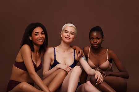 Three women with different skin color sitting together against brown background and smiling
