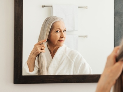 Aged woman with long gray hair looking at a mirror in the bathroom