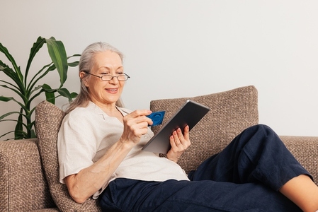 Smiling aged woman with a credit card  Mature female in eyeglasses holding a digital tablet while lying on a sofa at home