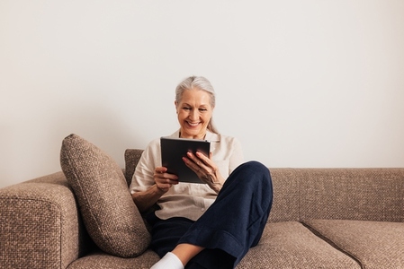 Adult cheerful female sitting on a sofa with digital tablet  Smiling senior woman holding a portable computer in the living room