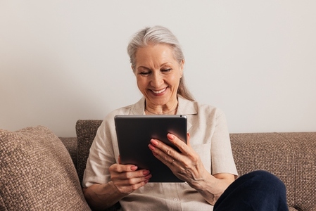Close up of a smiling senior woman with grey hair holding digital tablet while sitting on couch
