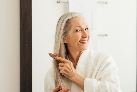 Cheerful woman combing her long hair with a wood comb in front of a bathroom mirror