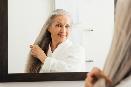 Mature woman combing her long gray hair in front of a mirror