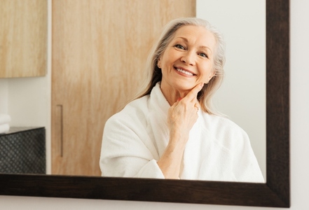 Portrait of an aged woman in a bathroom  Smiling woman with grey hair looking at her reflection in a mirror