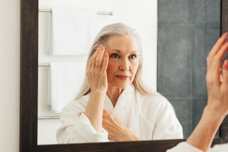 Aged woman looking at a mirror and touching her face  Female with grey hair wearing bathrobe standing in a bathroom