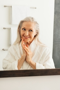 Portrait of an aged woman with folded hands in front of a bathroom mirror