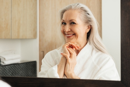 Aged woman admires her reflection in a bathroom mirror  Smiling senior female in a bathrobe