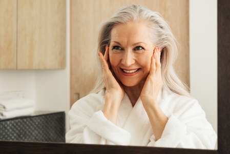 Smiling woman with grey hair massaging her face with her fingers  Aged female looking at her reflection in a mirror in the morning