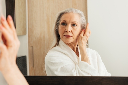 Smiling aged woman in bathrobe touching her face with fingers in bathroom