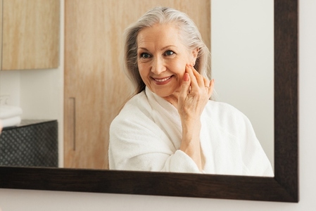 Mature woman with long grey hair looking at a mirror in a bathroom  Senior female in bathrobe adjusting her hair in front of a mirror
