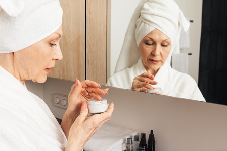 Senior woman with a wrapped towel on a head holding a cream  Aged female preparing to apply a moisturizer in bathroom