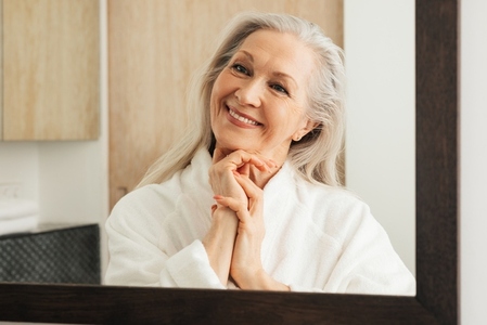 Cheerful senior woman with grey hair looking at mirror with folded hands