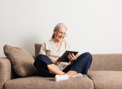 Cheerful aged female in glasses relaxing on a couch holds a digital tablet