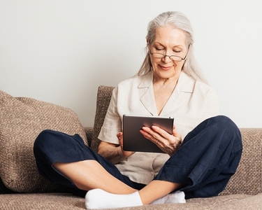 Senior woman with grey hair wearing glasses sitting with crossed legs on a couch using a digital tablet