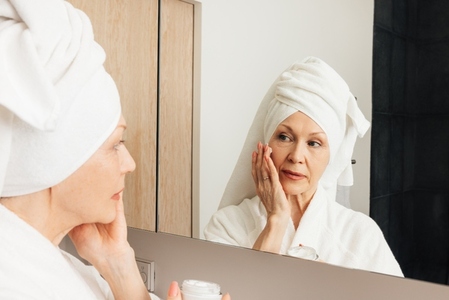 Senior woman with wrapped towel on her head applying cream on a face  Aged female doing morning routine in front of a bathroom mirror