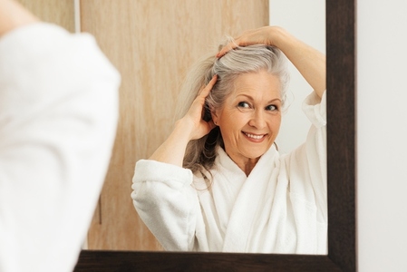 Smiling aged female making a new hairstyle by hands in front of a mirror