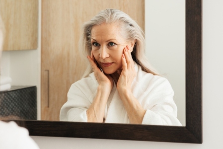 Senior woman in a bathrobe looking at a mirror and touching the face