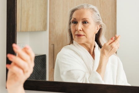 Portrait of an aged woman with grey hair looking at the mirror while standing in a bathroom